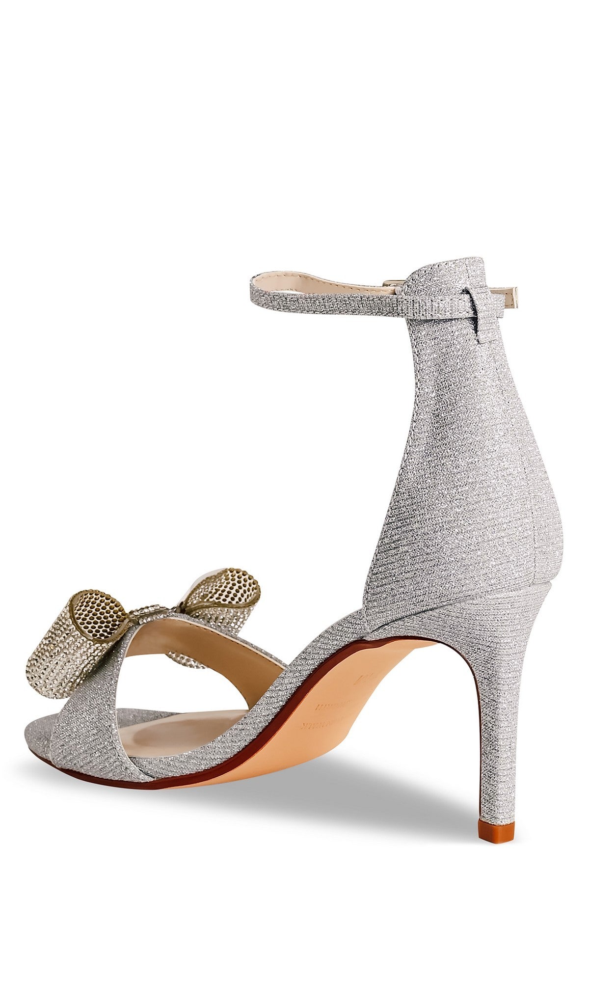 The Esme Silver Prom Shoes (Touch Ups 4744) feature a high heel, ankle strap stiletto, and a decorative bow on the front, displayed from a rear side angle.